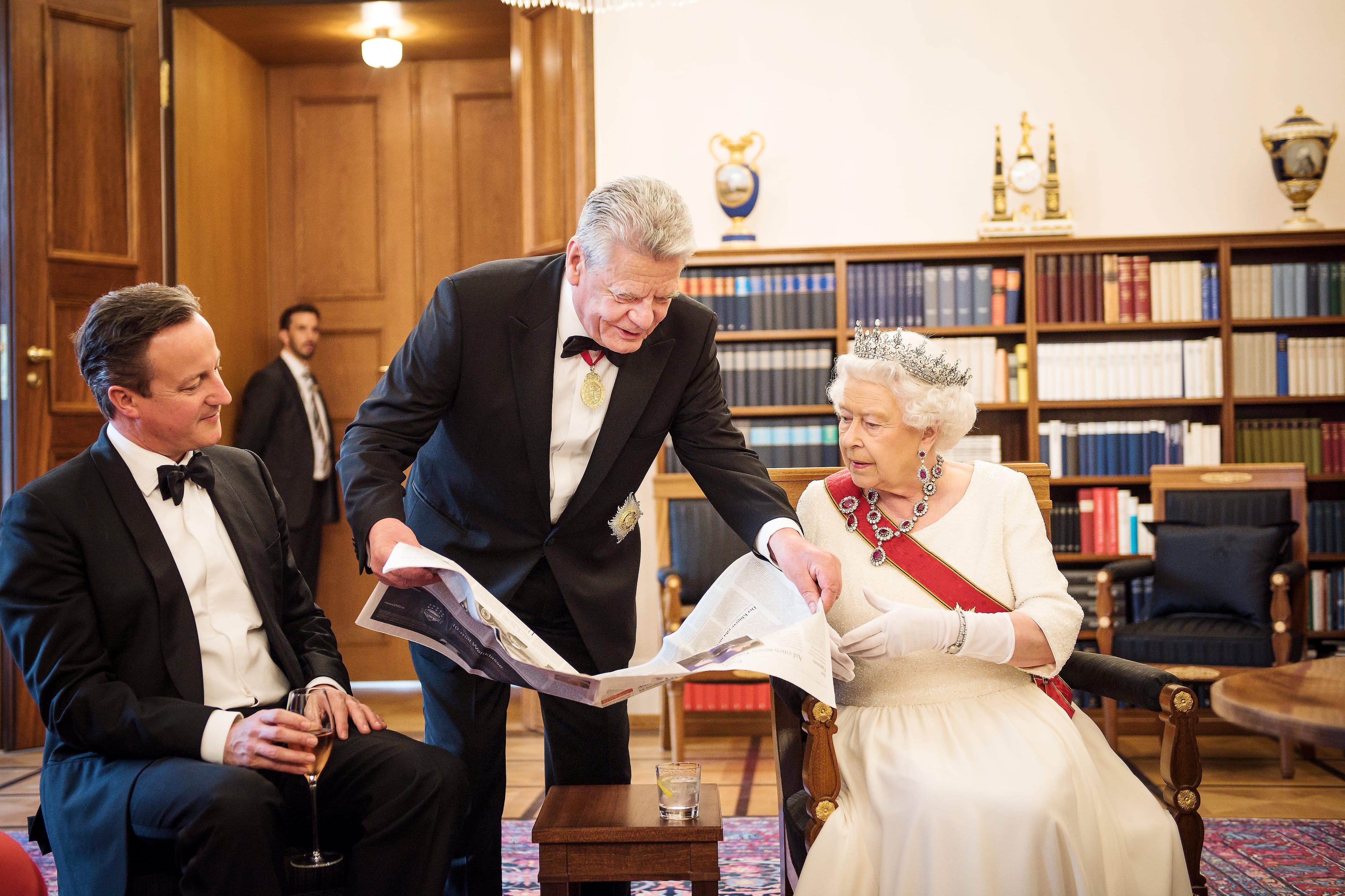 Bundespräsident Joachim Gauck, Queen Elisabeth II und der damalige britische Premierminister David Cameron betrachten bei einigen kurzen, gemeinsamen Momenten im Amtszimmer des Bundespräsidenten vor Beginn des Staatsbanketts, eine Seite aus der FAZ von 1989